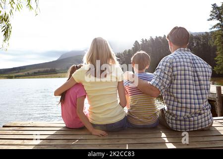 Bindung über natürliche Schönheit. Rückansicht einer Familie, die auf einem Pier auf einem See auf dem Land sitzt. Stockfoto