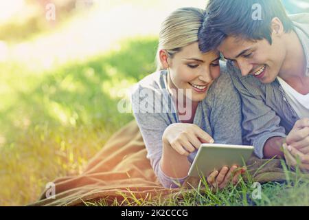 Im Garten stöbern. Aufnahme eines glücklichen jungen Paares, das auf dem Gras liegt und zusammen ein digitales Tablet verwendet. Stockfoto