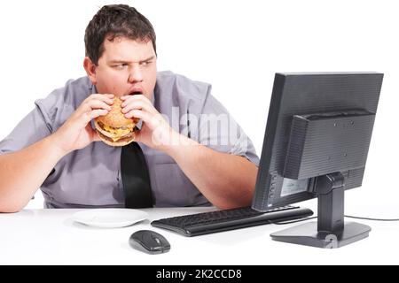 Hör auf, mir gesunde Dinge zu zeigen. Ein junger Mann isst sein Mittagessen an seinem Schreibtisch bei der Arbeit, während er mit dem Mund auf seinen Monitor starrt - ungesunde Essgewohnheiten. Stockfoto