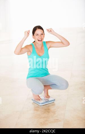 Ich habe mein Zielgewicht erreicht. Porträt einer attraktiven jungen Frau, die auf einer Skala hockend jubelt. Stockfoto