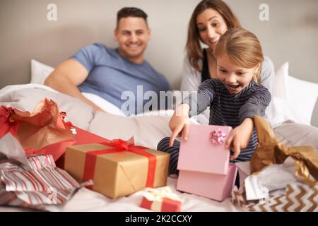 Oh wow, danke Mama und Papa. Aufnahme eines kleinen Mädchens, das Geschenke im Bett öffnet, während ihre Eltern zusehen. Stockfoto