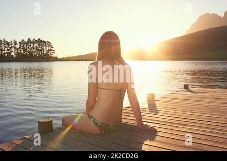 Der See bei Sonnenuntergang. Aufnahme einer jungen Frau in einem Bikini, die bei Sonnenuntergang auf einem Dock an einem See sitzt. Stockfoto
