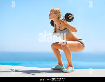 Heben Sie mit den Beinen. Ganzkörperaufnahme einer attraktiven jungen Frau, die draußen Kniebeugen mit einer Langhantel macht. Stockfoto