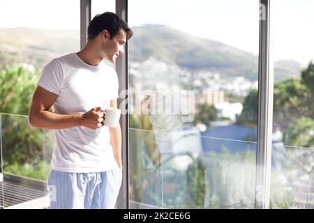 Beim Aufwachen gab es frischen Kaffee und eine tolle Aussicht. Eine kleine Aufnahme eines hübschen jungen Mannes, der Kaffee trinkt, während er an seinem Schlafzimmerfenster steht. Stockfoto