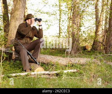 Wildtierjagd erfordert Geduld. Ein Wildhüter, der durch sein Fernglas schaut, während er auf einem Baumstamm sitzt. Stockfoto