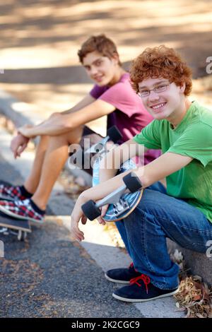 Das ist besser als drinnen zu sitzen. Porträt zweier junger Skateboarder, die an einer Straße sitzen. Stockfoto