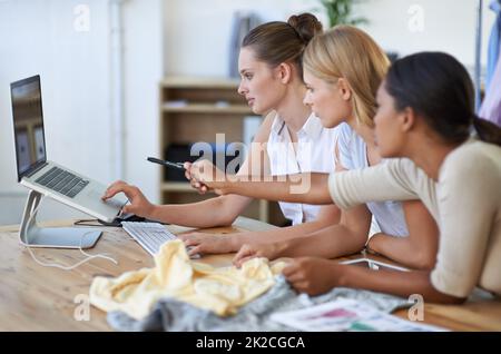 Lasst uns dieses Muster überprüfen... Drei junge Modedesigner diskutieren und zeigen auf einen Laptop. Stockfoto