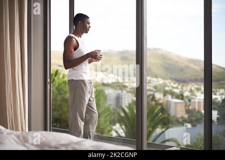 Es gibt nichts Schöneres als eine frische Tasse Kaffee, um Sie auf dem Weg zur Entspannung zu bringen. Aufnahme eines gutaussehenden Mannes, der beim Kaffeetrinken aus dem Fenster schaut. Stockfoto