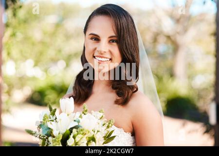 Heute werde ich den Knoten binden. Beschnittenes Porträt einer schönen jungen Braut, die lächelt, während sie einen Blumenstrauß an ihrem Hochzeitstag hält. Stockfoto