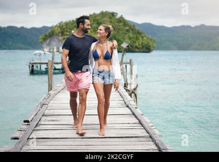 Ich liebe es, wie er mich ansieht. In voller Länge ein glückliches junges Paar, das während eines gemeinsamen Urlaubs Arm in Arm eine Promenade entlang läuft. Stockfoto