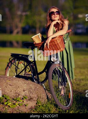 Genießen Sie die Ruhe der Natur. Aufnahme einer schönen jungen Frau, die sich im Park neben ihrem altmodischen Fahrrad entspannt. Stockfoto