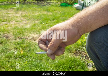 Nahaufnahme einer Hand, die eine Zigarette raucht, auf einem Grashintergrund, draußen, Raucherentwöhnungskonzept, Platz für Text links Stockfoto