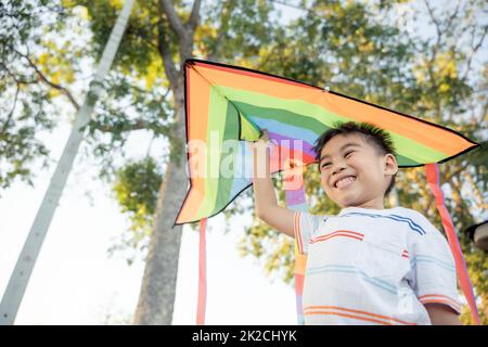 Asiatischer glücklicher Kinderjunge mit einem Drachen, der im Park weiterfliegt Stockfoto