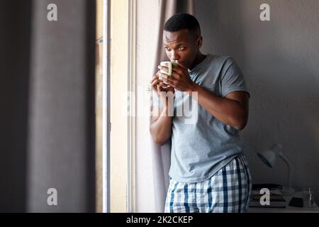 Es gibt nichts Schöneres als Kaffee, um den Tag zu überstehen. Gekürzte Aufnahme eines gutaussehenden jungen Mannes, der in seinem Schlafzimmer steht und nachdenklich aussieht, während er eine Tasse Kaffee genießt. Stockfoto