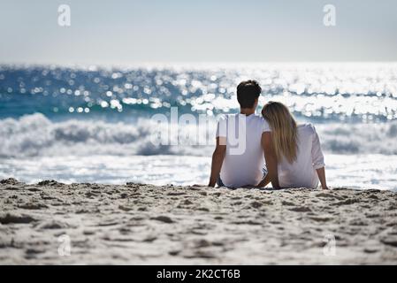 Über ihre Liebe nachdenken. Ein junges Paar sitzt zusammen am Strand und bewundert die schöne Aussicht. Stockfoto