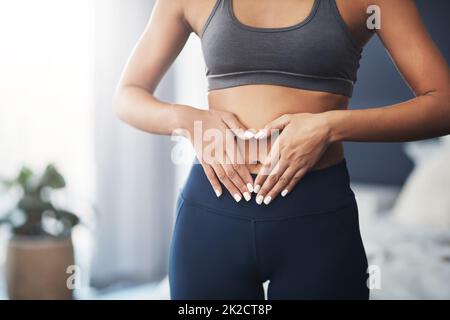 Du musst deinem Körper Liebe zeigen. Ausgeschnittene Aufnahme einer nicht erkennbaren jungen Frau, die in ihrem Schlafzimmer eine Herzform auf ihrem Bauch formt. Stockfoto