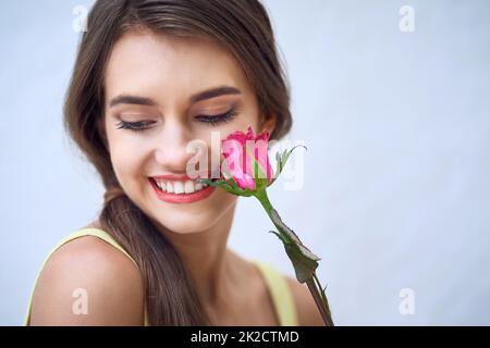 Zwei Naturschönheiten. Studio Nahaufnahme einer fröhlichen jungen Frau, die eine rosa Rose neben ihrem Gesicht hält, während sie vor einem grauen Hintergrund steht. Stockfoto