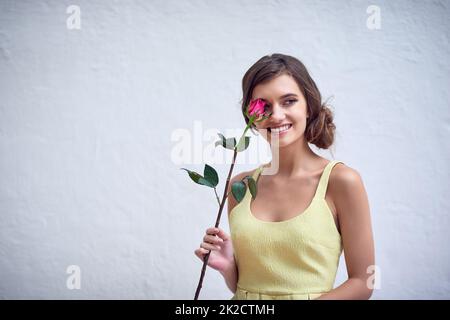 Shes eine Blume Art Mädchen. Studio-Aufnahme einer fröhlichen jungen Frau, die eine rosa Rose neben ihrem Gesicht hält, während sie vor einem grauen Hintergrund steht. Stockfoto