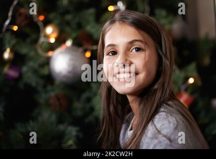 Es ist eine fröhliche Zeit des Jahres. Aufnahme eines kleinen Mädchens, das an einem weihnachtsbaum sitzt. Stockfoto