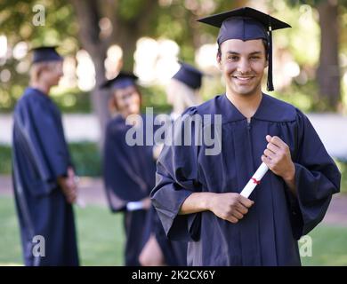 Begeistert von seiner Zukunft. Porträt einer lächelnden Absolventin, die sein Diplom mit ihren ehemaligen Absolventen im Hintergrund hält. Stockfoto