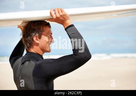 Es ist nur ich und mein Surfbrett. Aufnahme eines lächelnden jungen Surfers am Strand, der sein Brett auf dem Kopf trug. Stockfoto