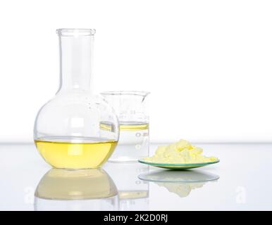 Chemische Inhaltsstoffe auf weißem Labortisch. Schwefelpulver in chemischem Uhrglas Platzierung neben Aluminiumchloridflüssigkeit, Öl und Alkohol im Becher. Seitenansicht Stockfoto