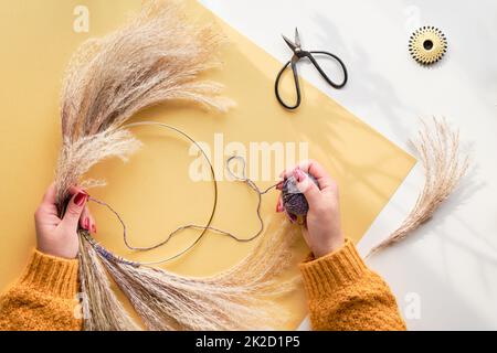 Hände, die getrockneten Blumenkranz aus trockenem Pampagras und Herbstblättern machen. Flach auf weiß gelbem Tisch, Sonnenlicht mit langen Schatten. Stockfoto