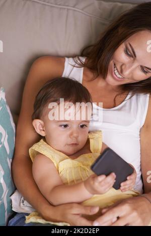 Es ist so neugierig. Ein Mädchen, das auf dem Schoß ihrer Mutter liegt und mit einem Handy spielt. Stockfoto