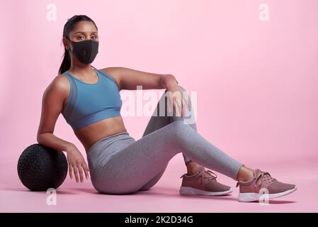 Ich bin entschlossen, meine Fitnessziele zu erreichen. Das lange Porträt einer attraktiven und sportlichen jungen Frau, die eine Maske trägt und mit einem Medizinball im Studio posiert, vor einem rosa Hintergrund. Stockfoto