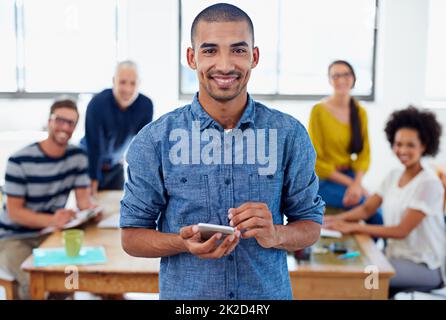 Mein Unternehmen ist technologiefreundlich. Porträt eines jungen Geschäftsmannes, der sein Telefon mit Mitarbeitern im Hintergrund hält. Stockfoto