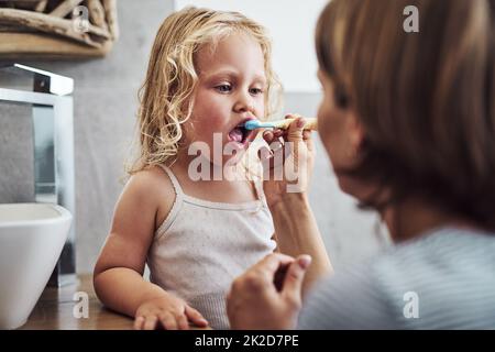 Mache ich es richtig Mama. Eine kurze Aufnahme eines entzückenden kleinen Mädchens, das beim Zähneputzen stand und Hilfe von ihrer Mutter bekam. Stockfoto