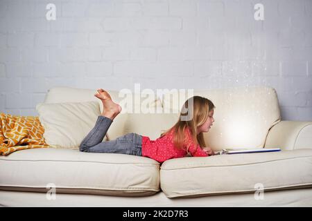 Es gibt so viel Magie beim Lesen. Aufnahme eines kleinen Mädchens, das ein Buch liest, während es auf einem Sofa liegt. Stockfoto