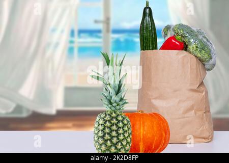 Nahaufnahme einer braunen Einkaufstasche aus Papier mit frischem Gemüse, einem Kürbis und einer Ananas auf dem Tisch über einem abstrakten, verschwommenen Meereshintergrund. Konzept der Stärkung des Immunsystems. Speicherplatz kopieren. Stockfoto