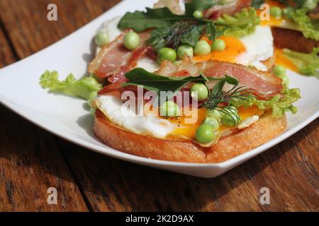 Bruschetta mit Rucola, chrispy Speck, Erbsen und pochiertem Ei auf einem weißen Teller serviert Stockfoto