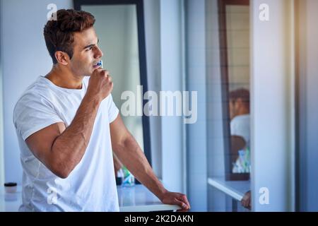 Tief in der morgendlichen Routinezone. Aufnahme eines hübschen jungen Mannes, der nachdenklich aussieht, während er sich zu Hause die Zähne putzt. Stockfoto