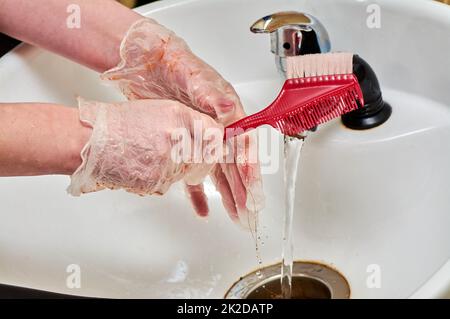 Frauenhände in Haarfärbehandschuhen waschen einen Kamm unter einem Wasserstrahl in einem weißen Waschbecken Stockfoto