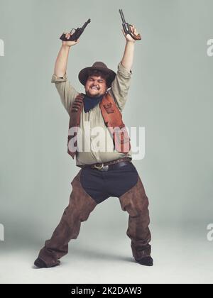 Er hörte, dass die Dorfleute einen neuen Cowboy brauchten Ein übergewichtiger Cowboy, der mit seinen Pistolen in der Luft ekstatisch aussieht. Stockfoto