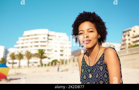 Wir fangen ein paar dringend benötigte Strahlen. Porträt einer attraktiven jungen Frau, die lässige Kleidung trägt, während sie allein am Strand sitzt und die Sonne genießt. Stockfoto