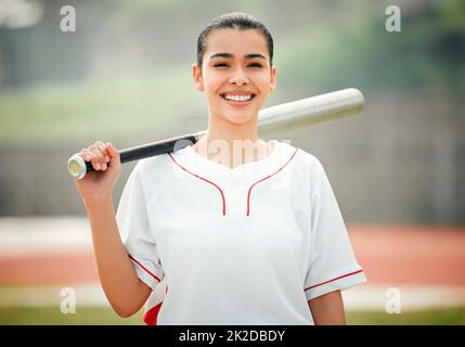 Teig aufschlagen. Beschnittenes Porträt einer attraktiven jungen Baseballspielerin, die einen Baseballschläger hält, während sie draußen steht. Stockfoto