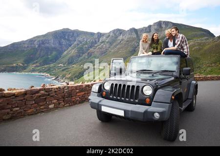 Halten Sie an, um die atemberaubende Aussicht zu erleben. Aufnahme einer Gruppe von Freunden, die sich auf dem Dach ihres LKWs mit einem bergigen Hintergrund entspannen. Stockfoto