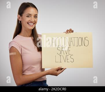 Es wird derzeit an COVID-19 geklebt. Aufnahme einer jungen Frau, die allein im Studio steht und ein Poster in der Hand hält, nachdem sie sich geimpft hat. Stockfoto