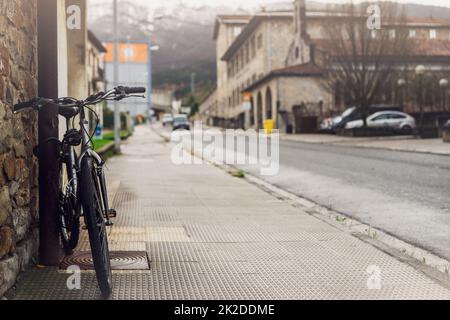 Das Fahrrad steht auf dem Gehweg in der Nähe der spanischen Stadtstraße. Das Fahrrad lehnt sich an die Stange neben dem alten Gebäude. Vorderansicht eines Fahrrads auf einem verschwommenen Gebäude, Autofahrt auf der Straße und Berghintergrund. Reisen in Europa. Stockfoto