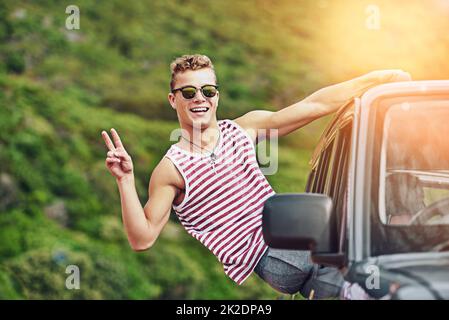 Nehmen Sie es einfach überall hin mit. Porträt eines jungen Mannes, der sich während einer Autofahrt aus dem Fenster eines Autos lehnt. Stockfoto