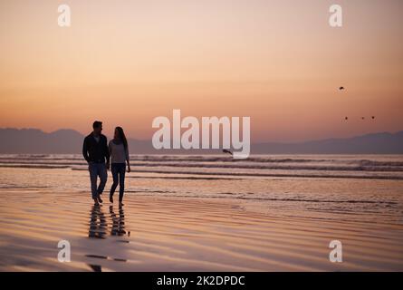 Es gibt nichts wie junge Liebe. Silhouette eines jungen Paares, das einen romantischen Spaziergang am Strand genießt. Stockfoto