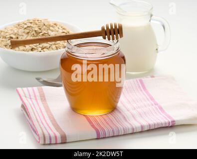 Honig in einem durchsichtigen Glas und ein Holzstab auf einem weißen Tisch, hinter einem Dekanter mit Milch und Haferflocken auf einem Teller Stockfoto