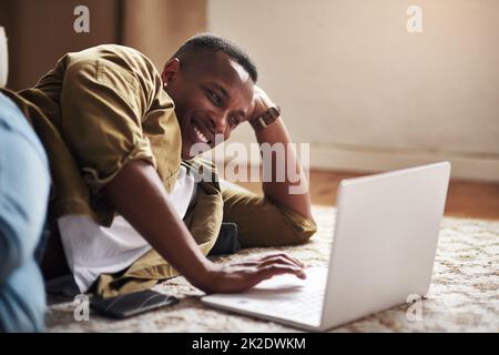 Mein Blog hat wirklich ein breites Publikum erreicht. Eine kleine Aufnahme eines hübschen jungen Mannes, der lächelt, während er zu Hause einen Laptop in seinem Wohnzimmer benutzt. Stockfoto