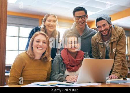 Es fühlt sich besser an, wenn man zusammen lernt. Porträt einer fröhlichen jungen Gruppe von Schülern, die mit einem Laptop für Prüfungen in einer Bibliothek arbeiten. Stockfoto