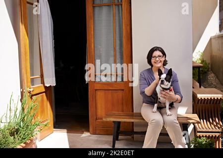 Alles, was man braucht, ist Liebe... Und einen Hund. Aufnahme einer Frau, die mit ihrem Hund auf einer Bank vor ihrem Haus sitzt. Stockfoto