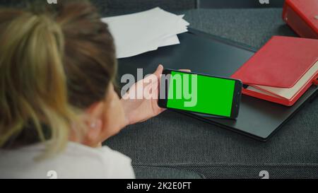 Frau sitzt auf einem Sofa mit Smartphone im horizontalen Modus mit nachgebildetem grünen Bildschirm Chrom-Key-Display. Stockfoto