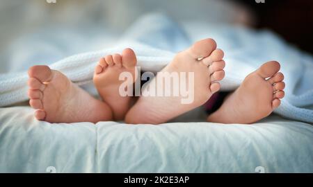 Früh ins Bett, früh zum Aufstehen. Zugeschnittenes Bild von den Füßen eines Elternteils und eines Kindes, die unter den Abdeckungen herausragen. Stockfoto